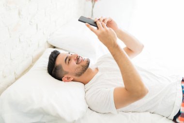 Gülümseyen iyi görünümlü adam yatakta akıllı telefonda komik videolar izliyor
