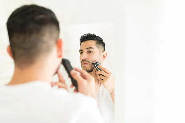 年轻人用电动剃须刀在浴室镜子前修剪胡子 — 图库照片
