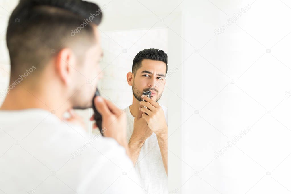 Good looking man looking in mirror while trimming his mustache in restroom