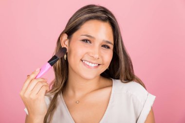 Young beautiful brunette applying blush and smiling while making eye contact
