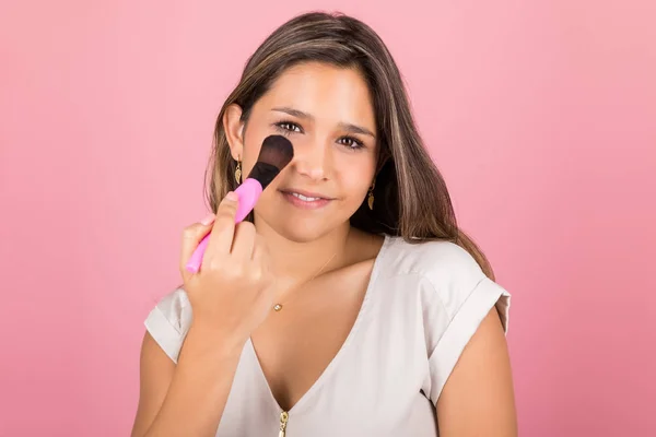 自信的拉丁妇女应用基础的脸与刷对粉红色背景 — 图库照片