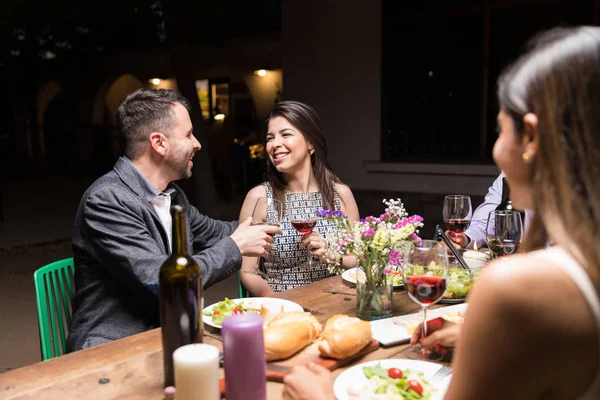 Smiling man and woman toasting red wine while enjoying dinner with friends at backyard party