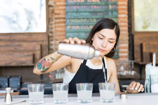 Female bartender pouring cocktail from shaker into glasses on bar counter