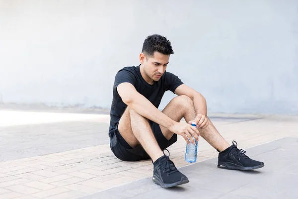 Tired male runner holding water bottle while sitting on footpath in city