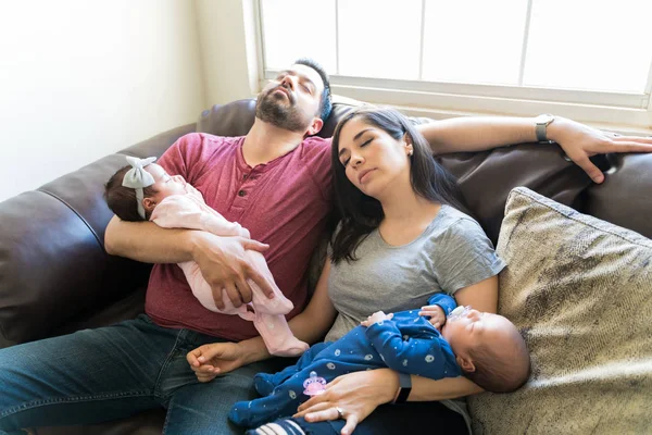 Tired Hispanic caring parents with newborn twins relaxing on sofa