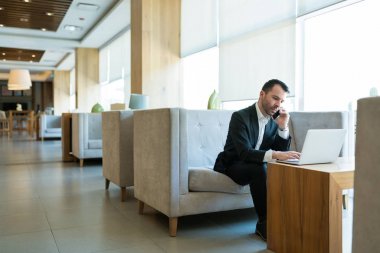 Cep telefonda konuşurken dizüstü bilgisayar kullanarak emin yakışıklı işadamı ve otel lobisinde kanepede oturuyor