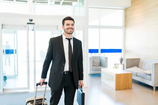 ホテルのロビーに到着するスーツケースを引っ張りながら歩く笑顔のイケメンエグゼクティブ — ストック写真