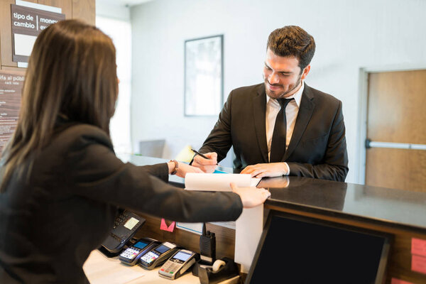 Smiling mid adult CEO filling booking form while standing with professional at counter in hotel reception