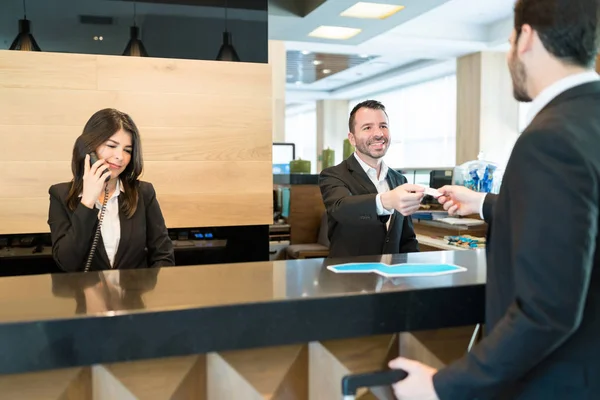 ホテルの受付エリアのカウンターで同僚が電話で話している間に立っている起業家にアクセスキーを与える笑顔のエグゼクティブ — ストック写真