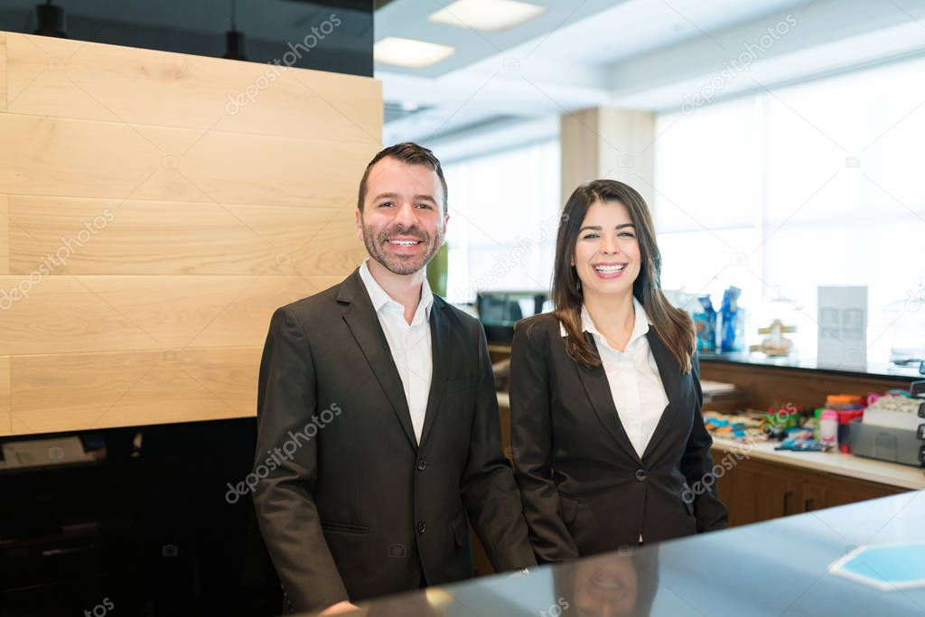 Portrait of Hispanic receptionist coworkers wearing suits while standing at desk in lobby at hotel