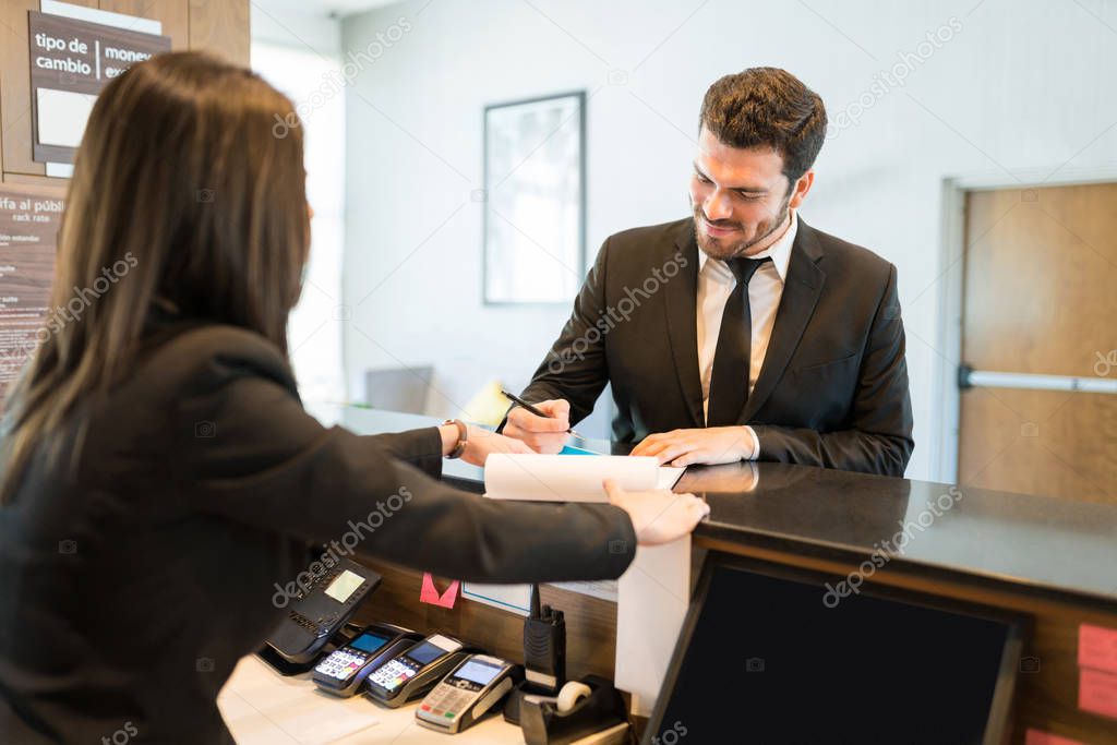 Smiling mid adult CEO filling booking form while standing with professional at counter in hotel reception
