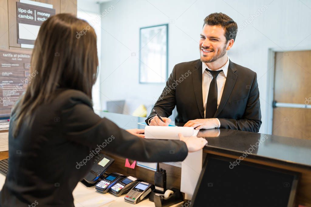 Happy Hispanic entrepreneur signing booking form while standing with clerk at front desk in hotel reception