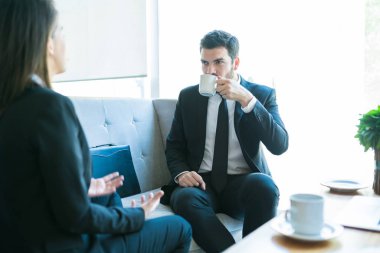 Seksi orta yetişkin profesyonel içme kahvesi, iş arkadaşını lobi toplantısında tartışmak için dinlerken