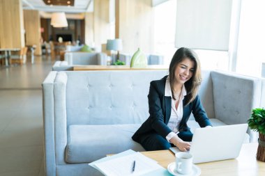 Otel lobisinde kanepede otururken laptop yazarak gülümseyen güzel kadın girişimci