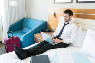 Otel odasında belgeler ile yatakta otururken meşgul erkek girişimci laptop üzerinde iş yapıyor