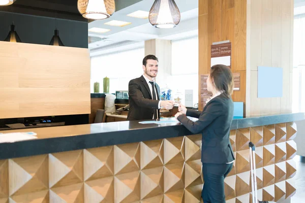 Smiling manager giving card key to female professional standing at front desk and welcoming to hotel
