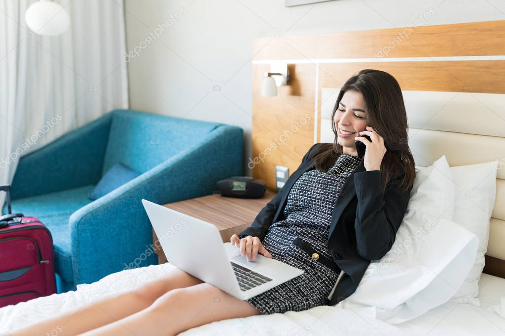 Smiling Hispanic businesswoman talking on smartphone while using laptop on bed in hotel room