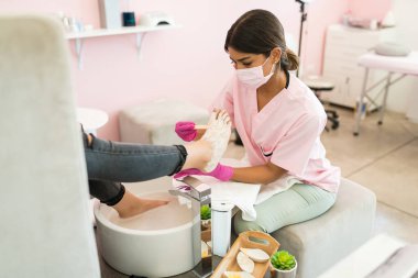 Spa tedavisi sırasında müşterinin ayağına krem uygularken maske takan pedikürcü 
