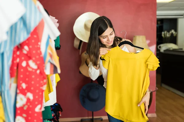 Beautiful young Latin woman looking at yellow top while standing by clothes rack in store