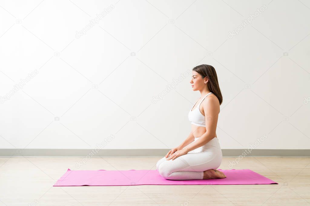 Side view of young attractive female performing Vajrasana on mat by wall in fitness studio
