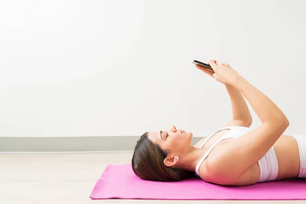 在健身室使用智能手机时 有吸引力的年轻女子躺在运动垫的侧视图 — 图库照片