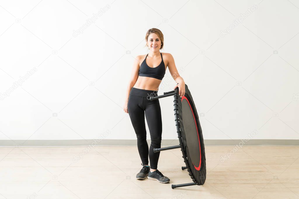 Full length portrait of beautiful young Caucasian woman standing with mini trampoline against white wall
