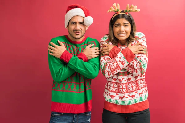 Portrait of boyfriend and girlfriend in ugly sweaters shivering during Christmas while standing against red background