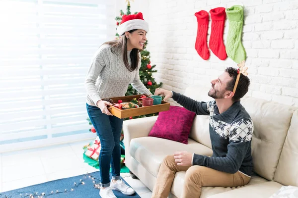 クリスマスシーズンに自宅のソファに座っている男性にスナックを提供する幸せな女性 — ストック写真