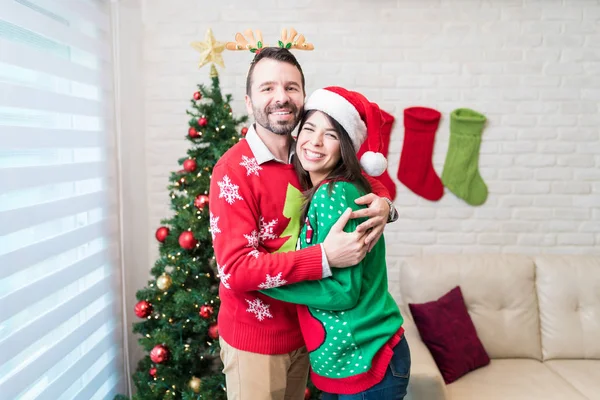 Loving cheerful couple in ugly sweater embracing while standing at decorated home during Christmas