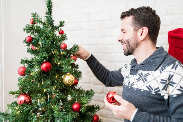 Smiling Caucasian man in sweater decorating tree with baubles at home during Christmas time