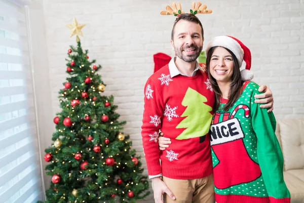 Smiling Caucasian couple in ugly sweater standing against Christmas tree at decorated home