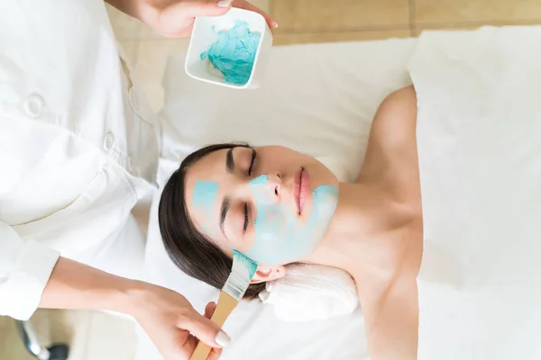 Pretty Latin female customer lying on bed while beautician applying facial mask with brush at spa