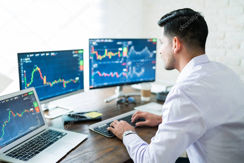 Hispanic young male stock trader typing on keyboard while using computer at home
