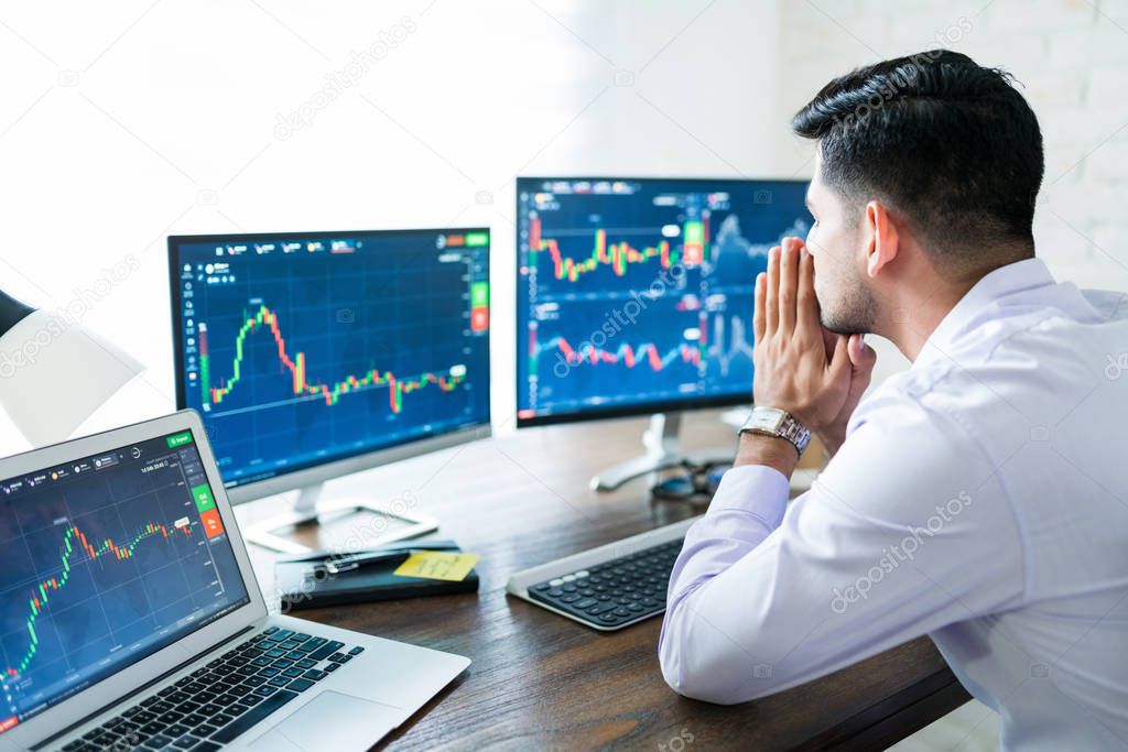 Young male stock broker praying for good fortune while trading online at desk from home