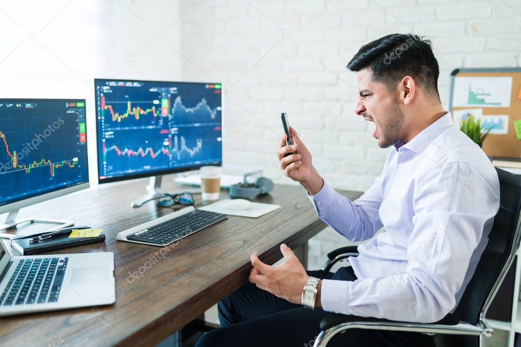 Frustrated Hispanic young freelance stock trader screaming while looking at mobile phone