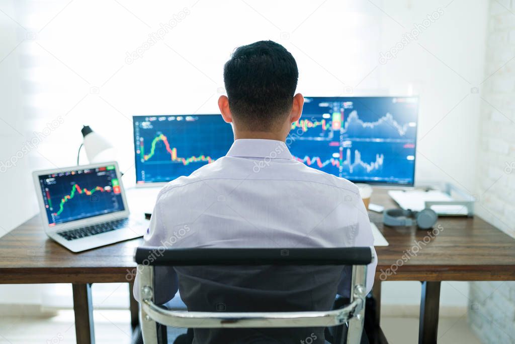 Rear view of young Latin male finance broker analyzing stock graphs while trading from home
