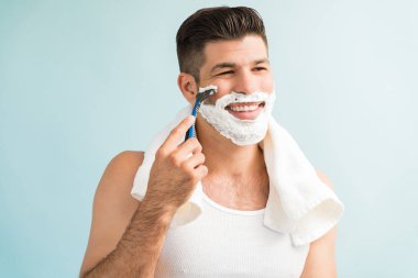 Genç Latin erkek jiletle tıraş olurken gülümsüyor ve turkuaz arka plana bakıyor.