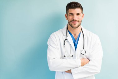 Kolları turkuaz arka planda çapraz dururken beyaz laboratuvar önlüğü giyen kendine güvenen çekici erkek doktor.