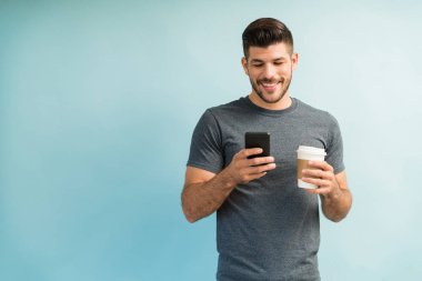 Gülümseyen çekici Latin adam. Akıllı telefondan mesaj atıyor. Elinde kahve fincanı tutarken arka planda duruyor.