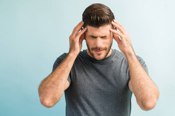 Young male Latin in discomfort suffering from headache while standing against plain background