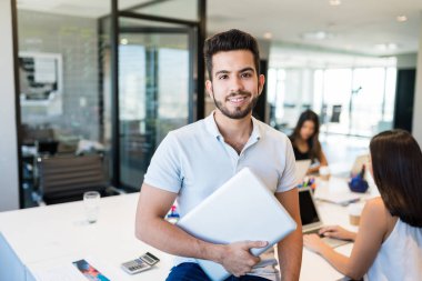 Sırıtan genç İspanyol iş adamı dizüstü bilgisayarla ofiste oturuyor.