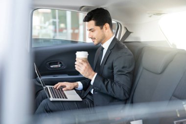 Takside dizüstü bilgisayar kullanırken kahve içen kendine güvenen Latin işadamı