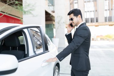 Kendine güvenen İspanyol işadamı araba kapısını açarken cep telefonuyla konuşuyor.