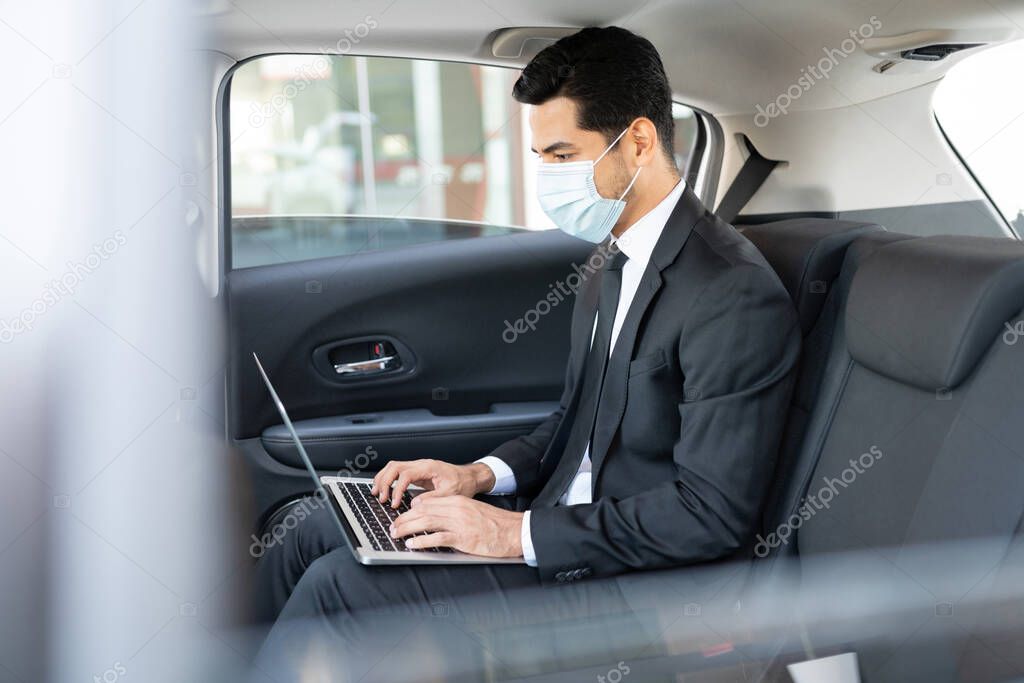 Young businessman wearing face mask while using laptop in taxi during coronavirus outbreak