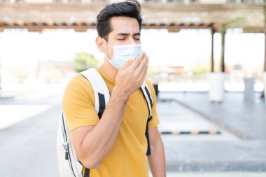 Latin genç adam COVID19 salgını sırasında şehirde maske takarken öksürüyor.