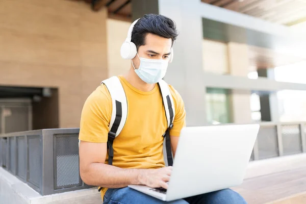 Latin young man in face mask listening music while using laptop in city during coronavirus crisis