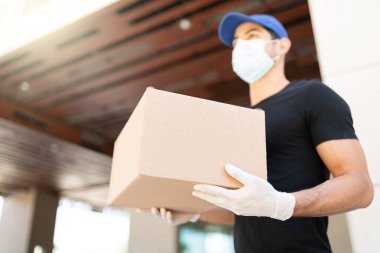 Young Latin delivery man wearing face mask while carrying parcel box during COVID19 crisis. Focus on package clipart