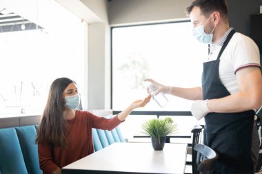 Lokantada masada oturan genç bir kadının elinde dezenfektan döken erkek personel.
