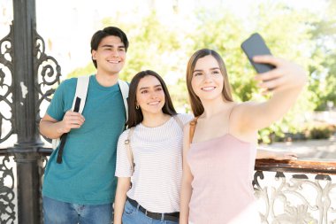 Genç bir kadın arkadaşlarıyla birlikte akıllı telefondan fotoğraf çekiyor ve dışarıda gülümsüyor.