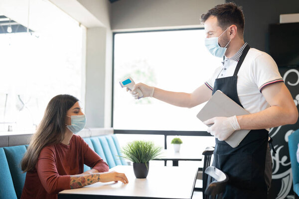 Staff checking body temperature of customer sitting at table through thermal scanner in cafe